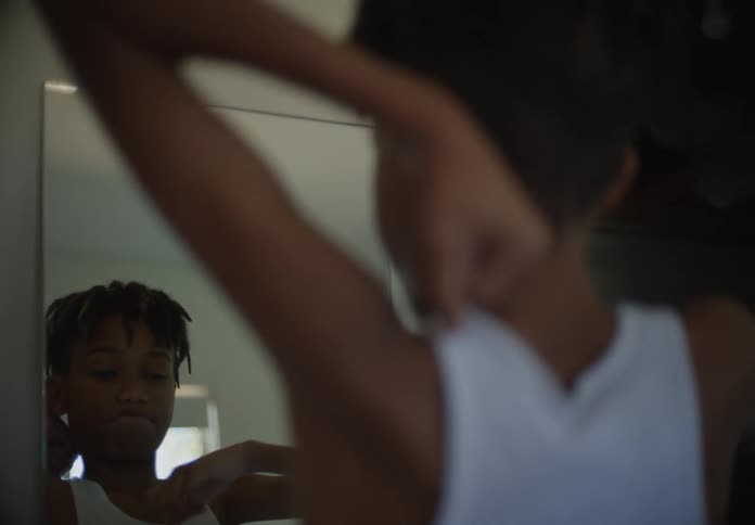Teenage boy with dreadlocks and white undershirt looks in a mirror in room