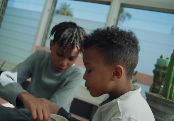 Two boys sitting together while one eats cereal out of a bowl and the other picks pieces out to eat