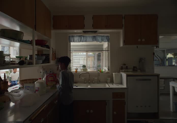 Youth pours milk onto cereal in kitchen while pass through shows glimpse of his younger brother