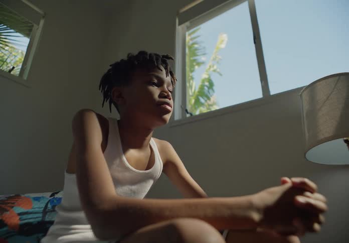 Boy with dreadlocks and white undershirt sits on bed in bedroom