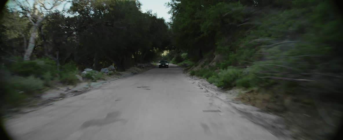 A black car travels quickly on a rural road surrounded by trees