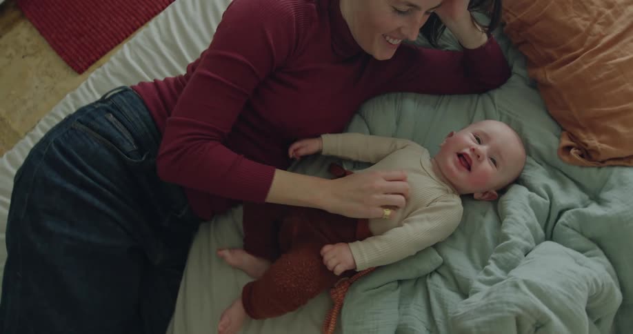A mother lays on the bed with her baby daughter as they play and smile