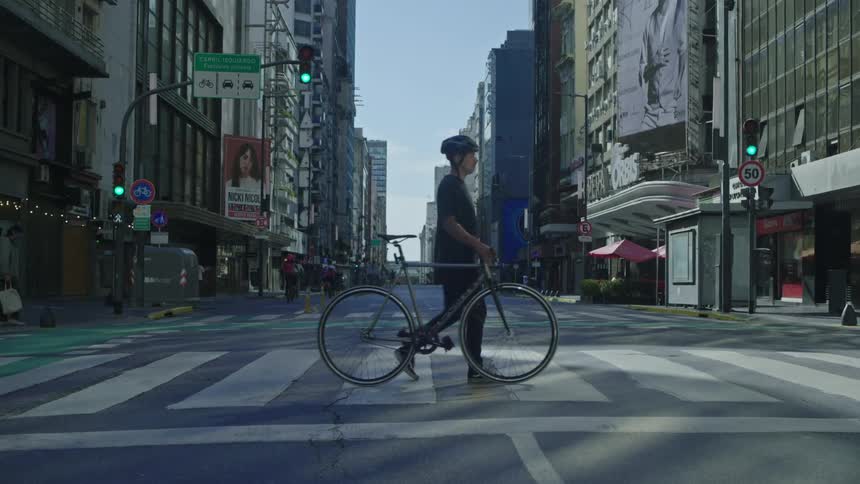 A man walks his bike across a city street as another rides down the street and people walk