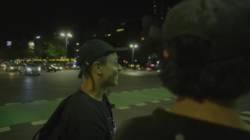 Two men stand on a busy street corner and talk and laugh as cars drive past