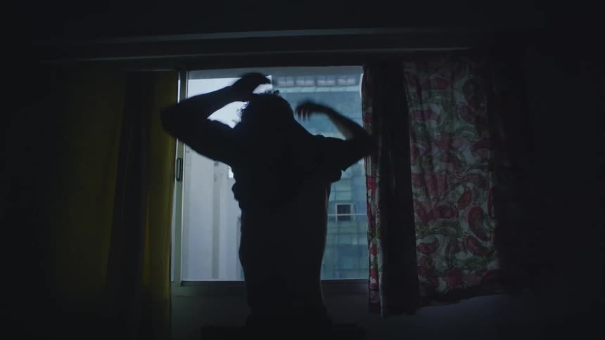 Man puts on shirt while standing near window in dim room