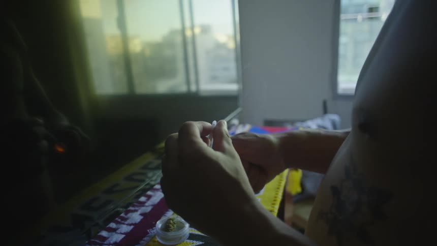 A man with his shirt off standing in front of a television lighting a rolled up blunt