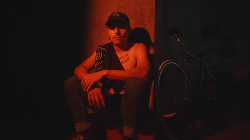 Shirtless man sitting on sidewalk under red lighting next to wall with bicycle next to him