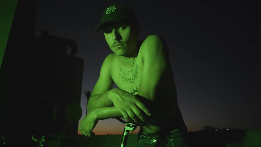 A man without a shirt leans on his bike at night in a green light