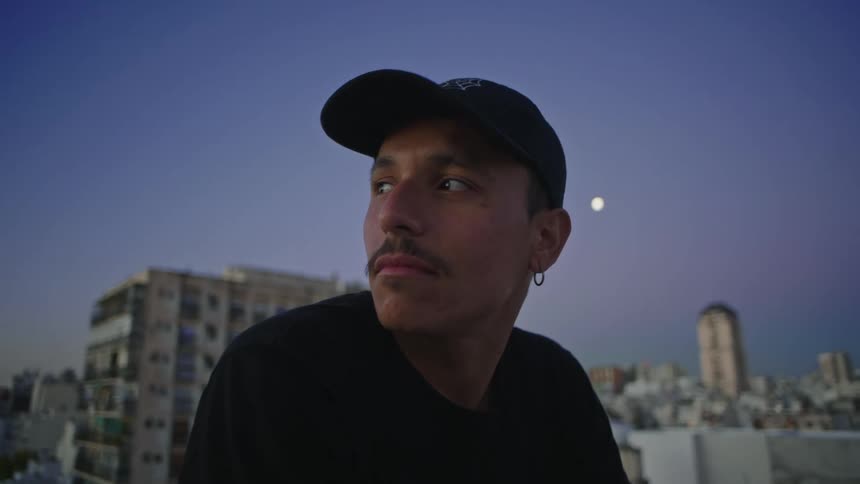 A man sitting with a black cap on outdoors with a city behind him