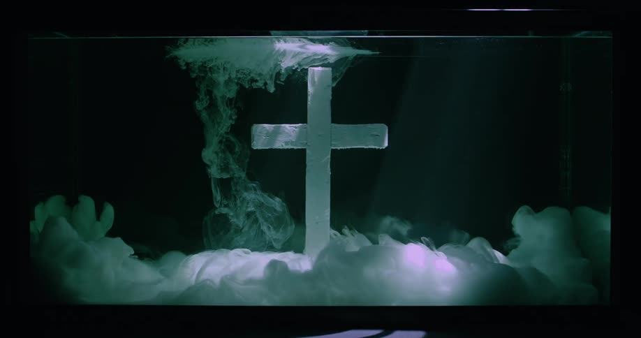 White cross submerged in water surrounded by clouds of white liquid ink under shifting lights