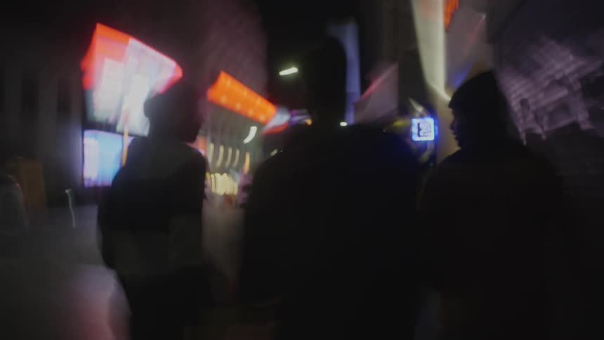 Men walk along sidewalk at night lit by bright neon signs