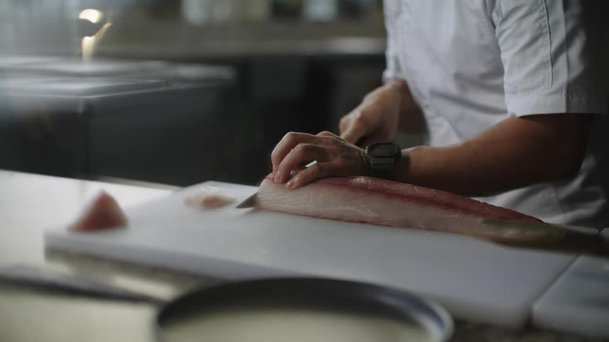 Chef uses a large sharp kitchen knife to slice a fish over the chopping board at a restaurant