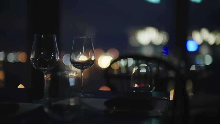 Wine glasses sit on table in restaurant a short distance from windows showing city lights at night