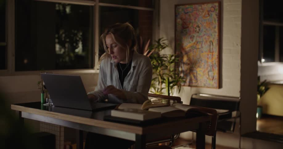 Young woman reads a book while walking to sit on her desk and use a laptop