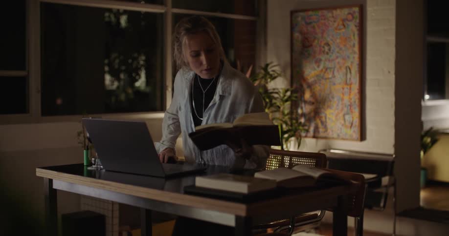 Young woman reads a book while walking towards her desk to use a laptop