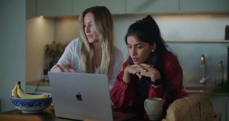 Two women in kitchen discuss contents on apple laptop screen