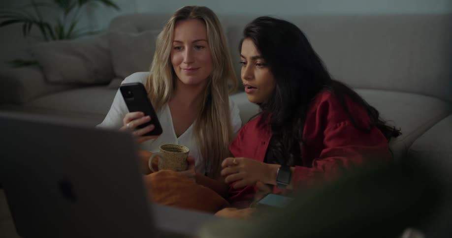 Two young women sitting between a couch and table talk about something shared on a cell phone