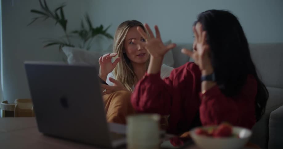 Two women sit on floor in living room at coffee table with fruit as they type on laptop and converse
