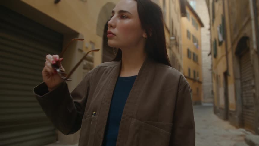 A dark haired woman walks down a corridor outdoors and puts her sunglasses on