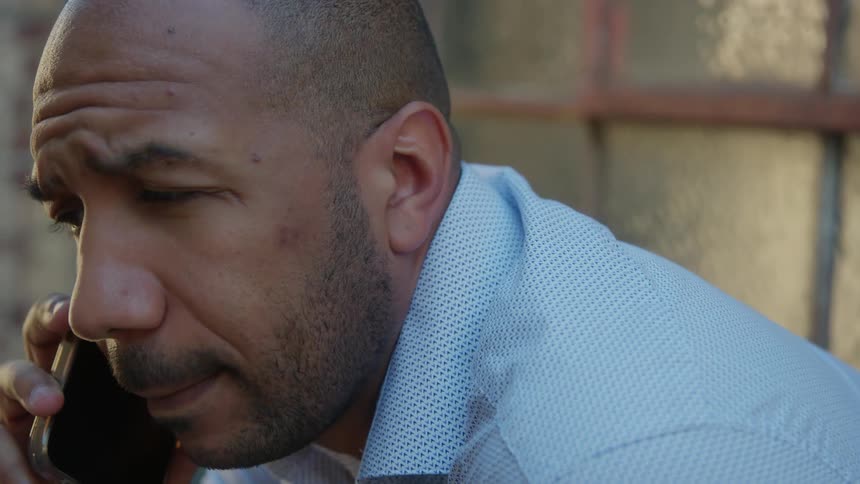 A bald man wearing a light blue shirt talks on his cell phone outside