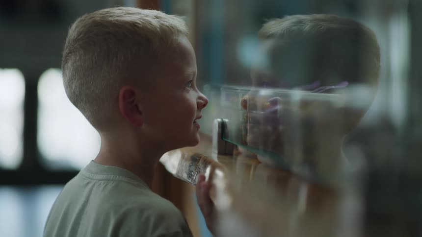 Close up of the profile of a blonde curious boy looking through a window in amazement