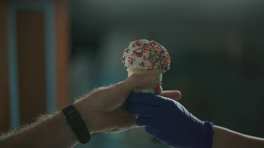 A person wearing a purple glove hands an ice cream cone with candy sprinkles to a customer
