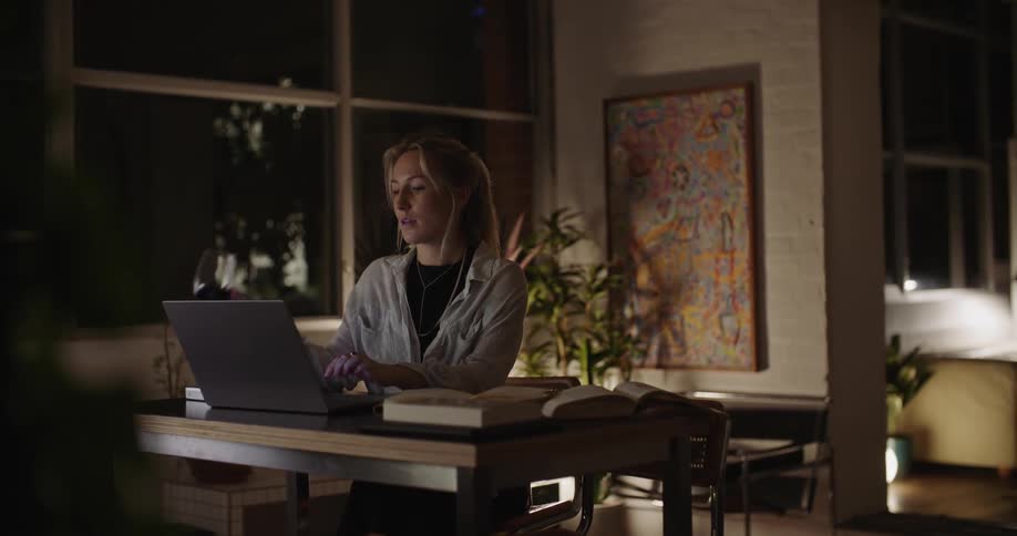 A woman is sitting at a table looking at a book and typing on a laptop computer