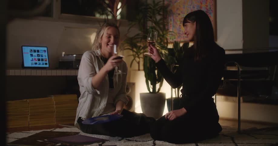 A pair of women clink their wine glasses before taking a drink while sitting together on the floor indoors at night