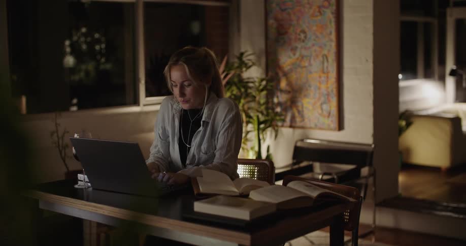 A woman is sitting at a table with a book as she types on her laptop computer
