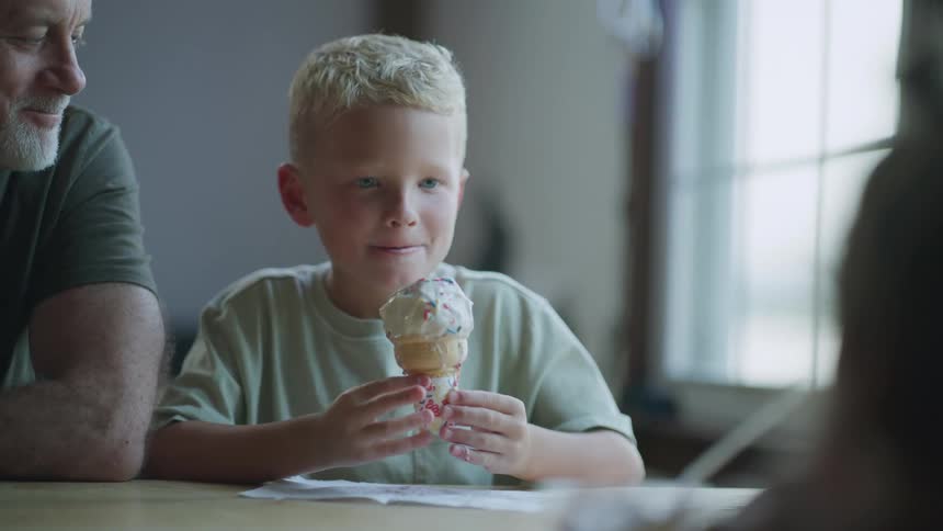 Grandfather sits at table smiling while his grandson eats an ice cream cone across from his granddaughter