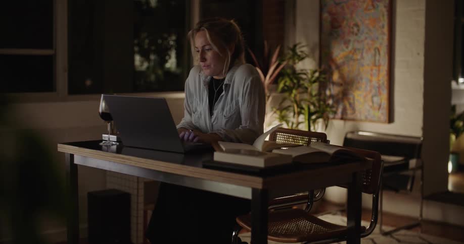 A woman walks to her laptop with wine and a book