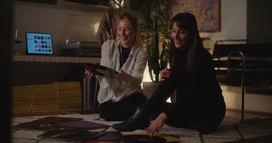 Two women are sitting on the floor discussing a record collection that is spread out in front of them