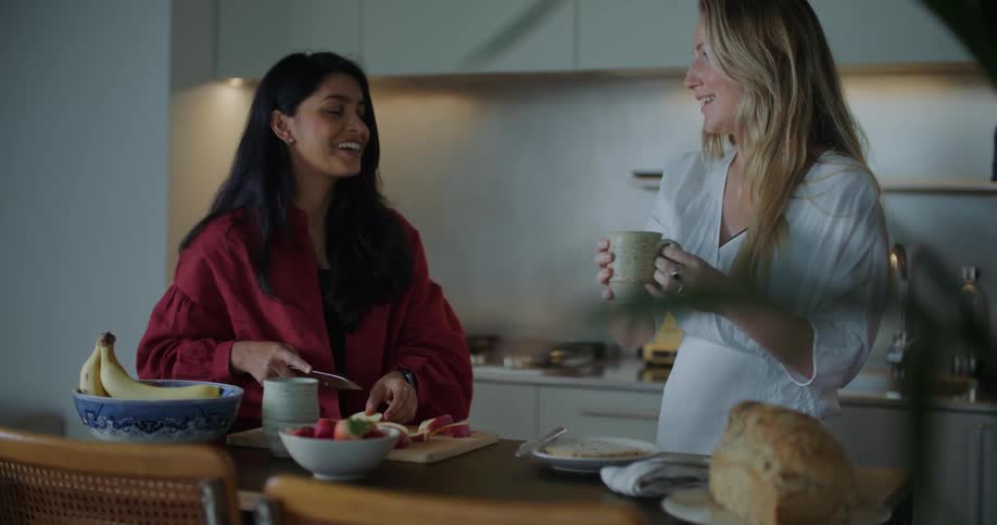 Two woman are talking and laughing in a kitchen while they make breakfast