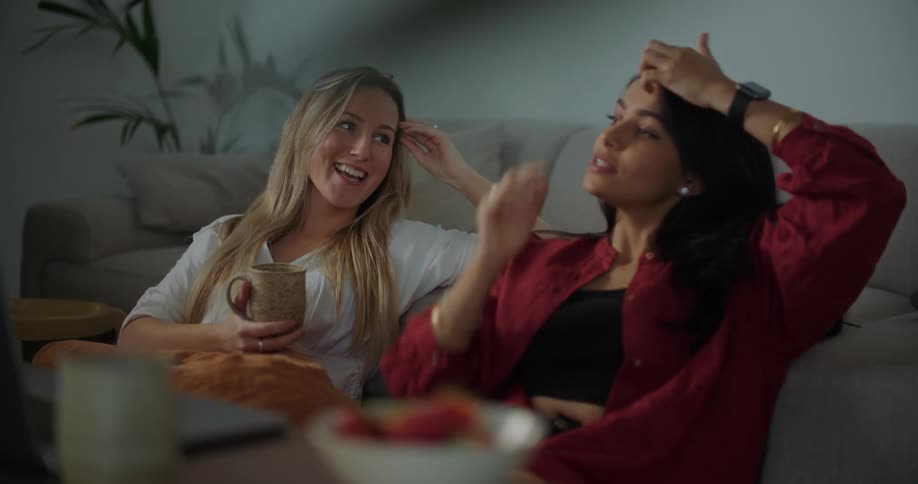 Blonde woman with white shirt and woman with dark hair and red shirt sits on the floor in the living room talking with each other