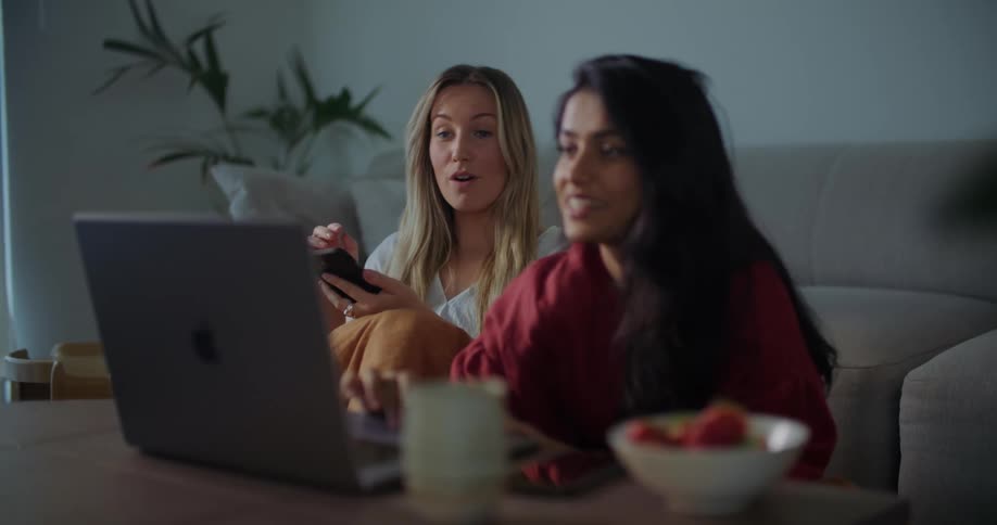 Two women are sitting on the floor in front of a table looking at a laptop computer and talking as one woman types as the other holds her phone