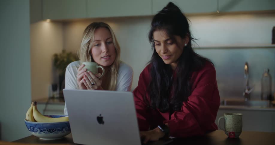 Two friends reading on a laptop screen and talking in the kitchen at home