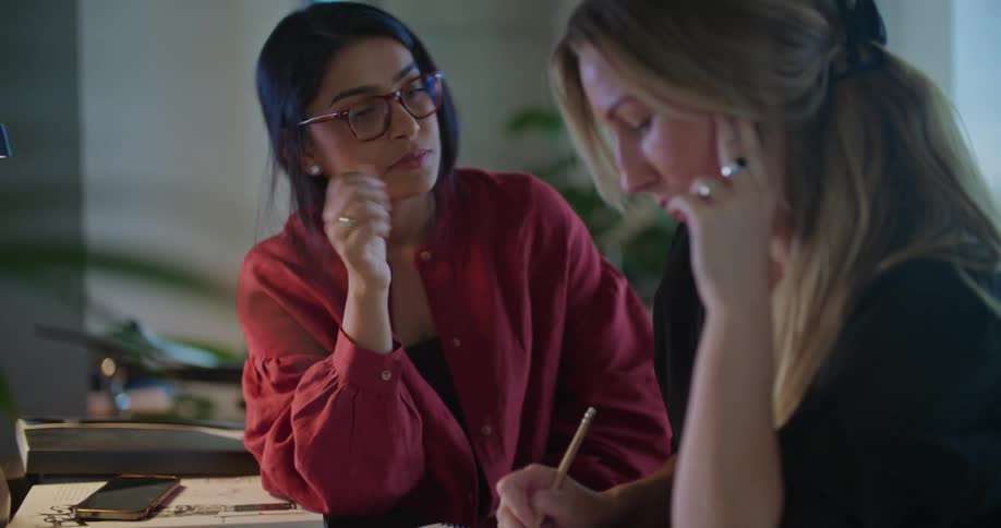 Two women sitting at an office desk near a laptop and cell phones work with paper and pencil