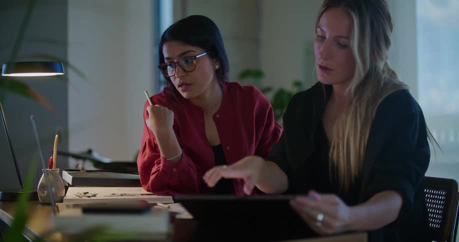 Two women sit at table discussing plans as they consult various computers