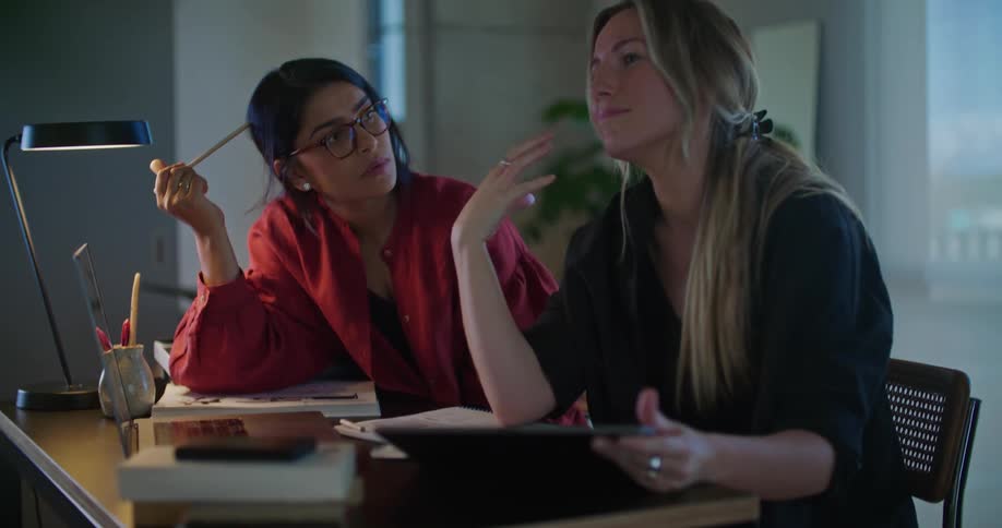 Two women are sitting at a table looking at a laptop computer and pointing to the screen as they talk and one woman is holding an open book