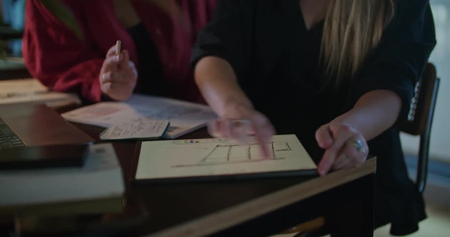 Two young women work at an office desk while one uses her fingers to draw on a tablet