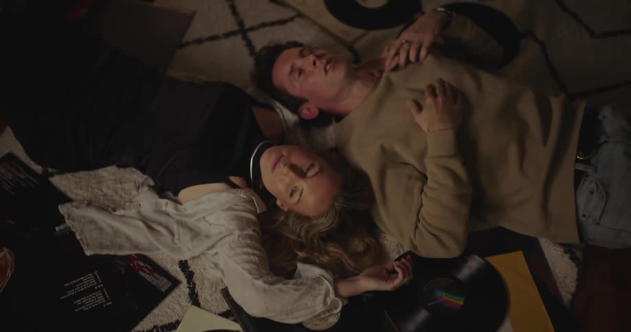 Man and woman lie on floor among vinyl records in opposite directions with their heads together as they talk