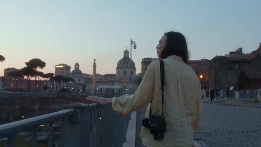 A woman with a camera walks near a railing in a city center that has big old buildings nearby