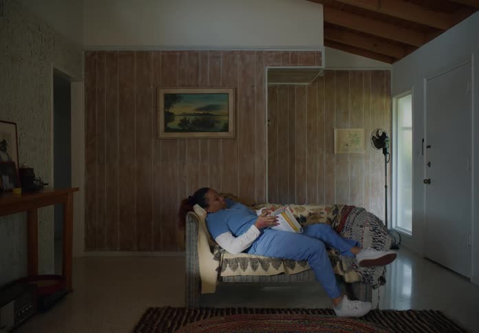 A nurse enters a living room and removes her jacket before relaxing on the sofa with a textbook