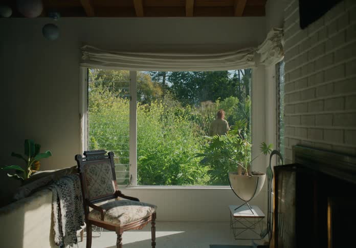 A woman walking around in her yard full of plants while her two sons watch her from the window inside