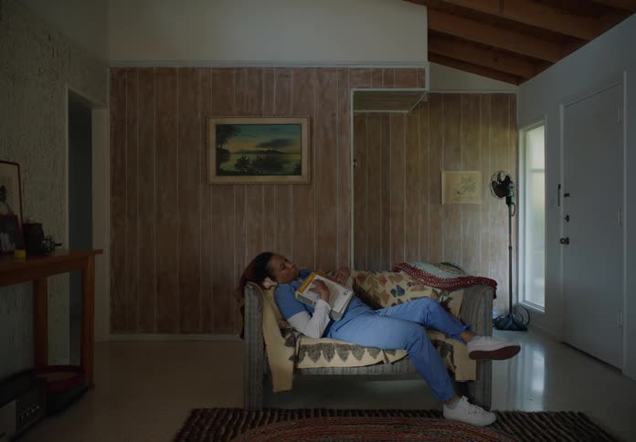 A nurse walks in to her home and picks up a book and lies on the couch and falls asleep and then her son comes in and covers her up and puts the book away
