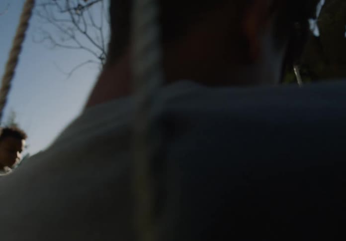A boy is sitting on a swing in his yard moving back and forth as his young brother walks by the tree
