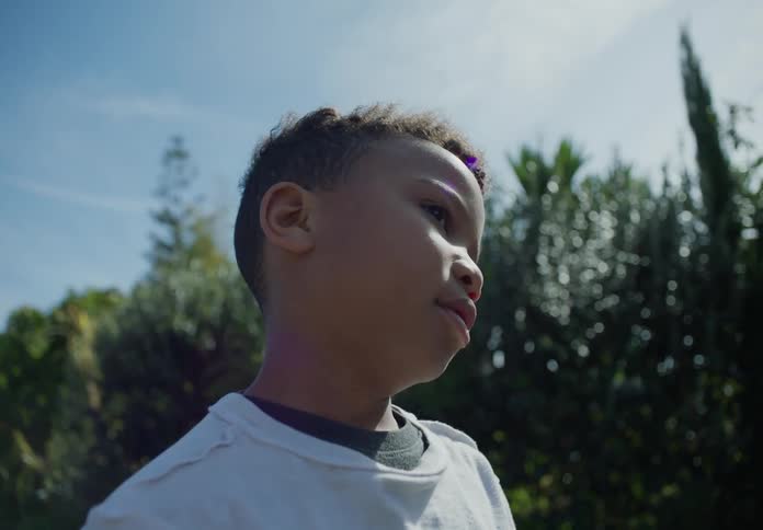 A young black boy stands outside near some trees on a sunny day