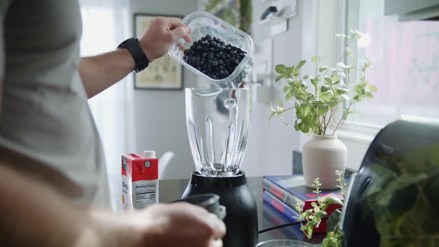 A man pours blueberries into a blender near a kitchen window