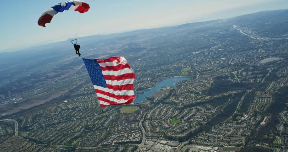 Person paraglides over town trailing a large fluttering american flag