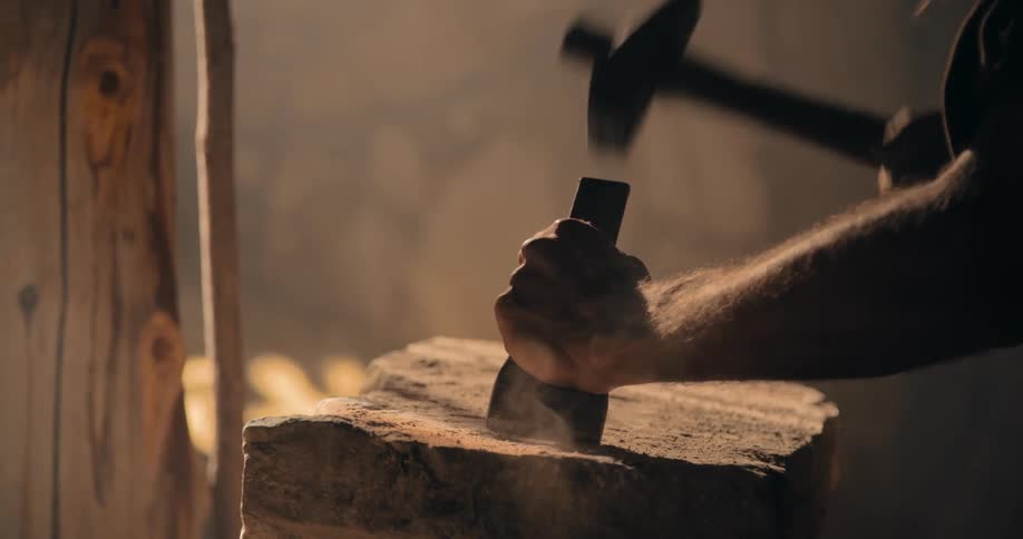 A man hammers a metal chisel into a piece of stone creating dust